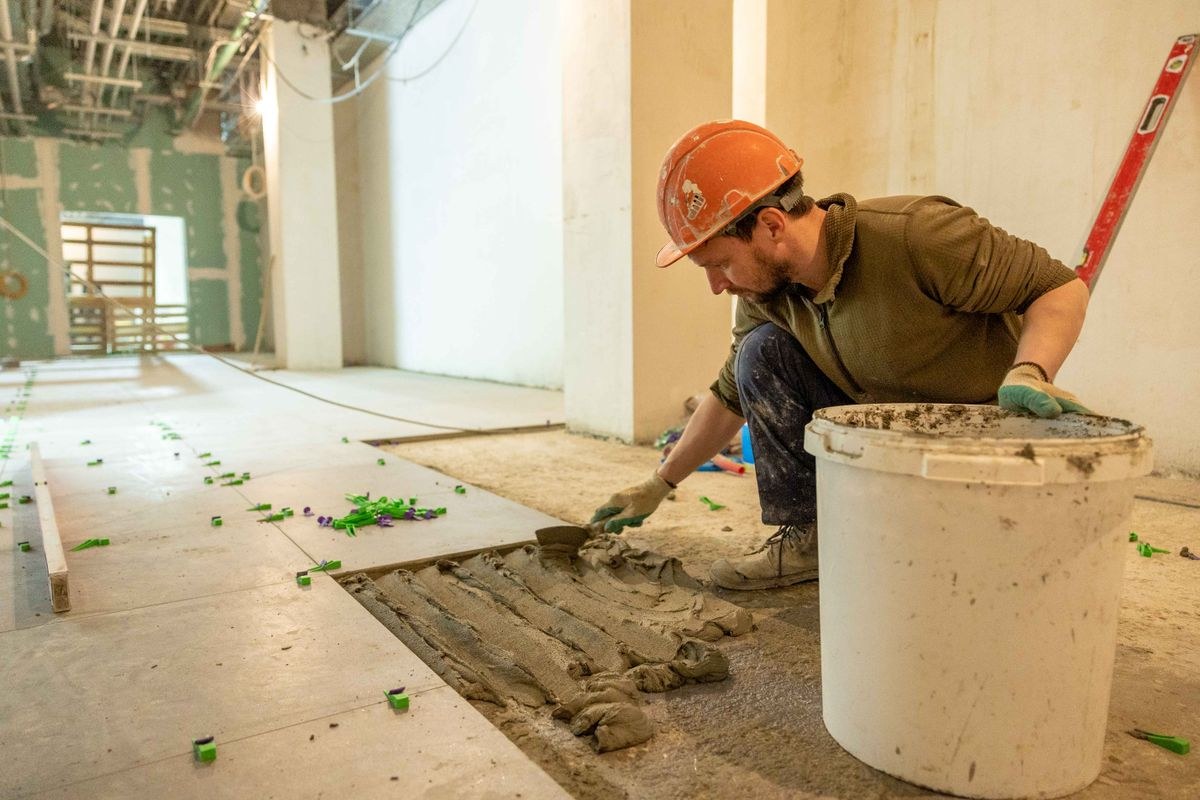 Tiler in an orange hard hat spreading notched adhesive on a screed floor beside newly laid large pale tiles held by green levelling clips